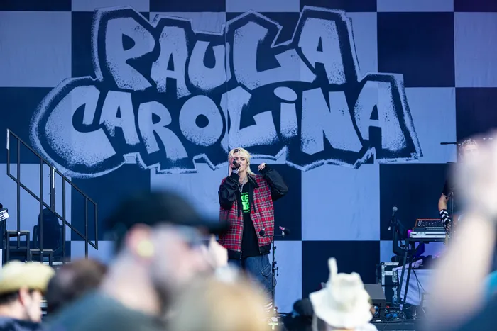 Paula Carolina singt mit einem Mikrofon vor einem großflächigen schwarz-weiß karierten Hintergrund mit ihrem Namen.