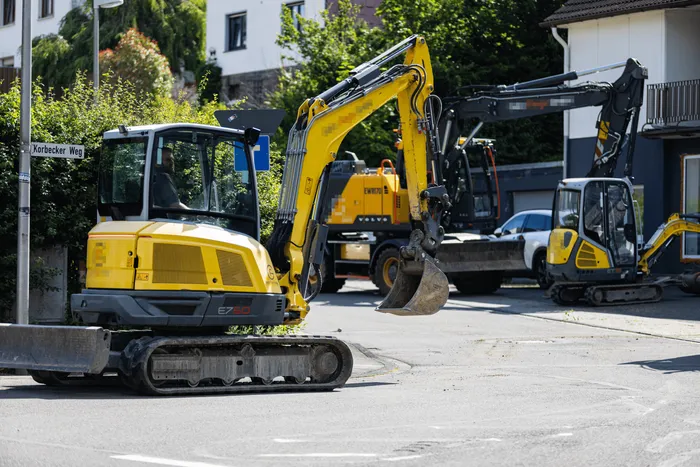 Gelbe Minibagger stehen auf einer Straße vor Häusern, während im Hintergrund ein weiterer Bagger zu sehen ist. Ein Straßenschild mit der Aufschrift „Korbecker Weg“ ist links im Bild zu erkennen.