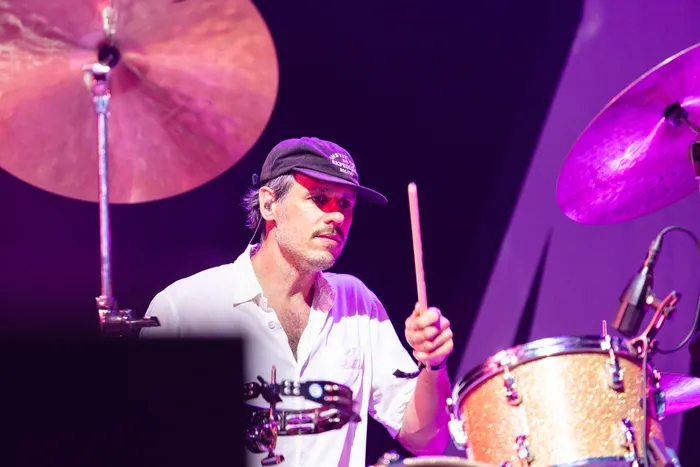 Schlagzeuger der Band Juli mit weißem Hemd und Cap spielt bei einem Auftritt unter violettem Bühnenlicht.