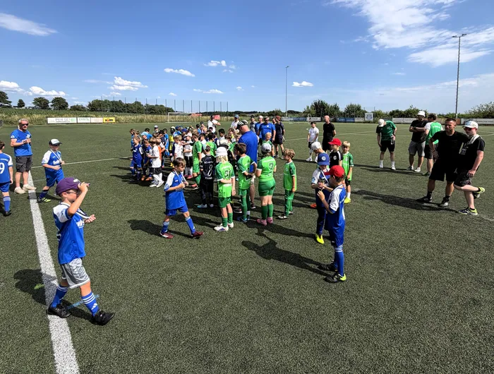 Eine Gruppe Jugendlicher in Fußballtrikots steht auf einem Kunstrasenplatz zusammen, umringt von Trainern und Betreuern. Im Hintergrund sind weitere Personen und ein Zaun zu sehen.