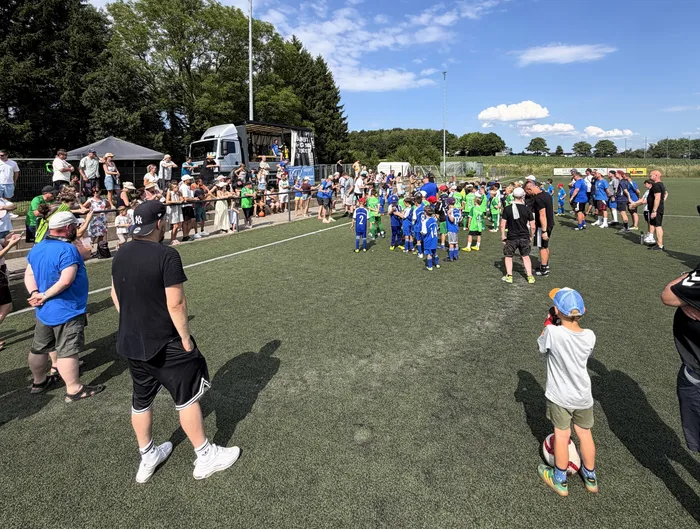 Fußballspieler in blauen und grünen Trikots stehen auf einem Rasenplatz, umgeben von Zuschauern und Betreuern. Im Hintergrund ein Mannschaftsbus und Bäume unter blauem Himmel.