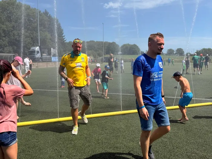 Zwei Männer gehen auf einem grünen Kunstrasenplatz, während Wasserfontänen um sie herum sprühen. Im Hintergrund sind weitere Personen und ein Parkplatz zu sehen.