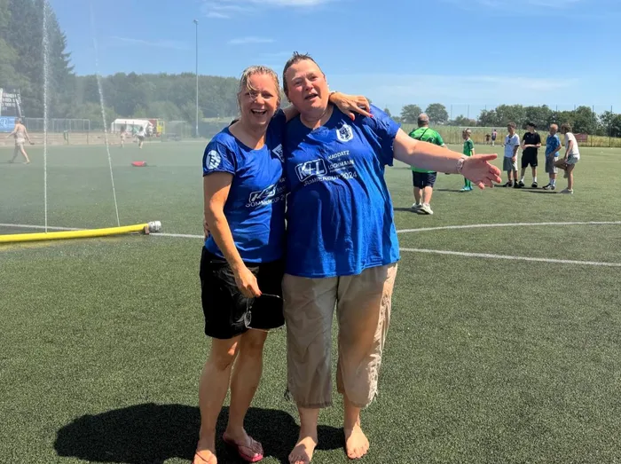 Zwei lächelnde Frauen in blauen Trikots stehen auf einem grünen Kunstrasenplatz, die eine hält eine Sprühflasche. Im Hintergrund sind weitere Personen und ein Fußballfeld zu sehen.