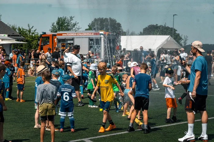 Fußballspieler in Vereinskleidung stehen auf einem grünen Rasenfeld, umgeben von Zuschauern und einem Feuerwehrauto im Hintergrund. Im Hintergrund ist ein weißes Zelt zu sehen.