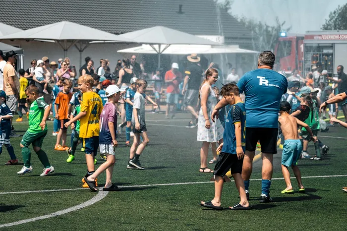 Kinder und Jugendliche spielen Fußball auf einem grünen Rasenplatz, während im Hintergrund Zuschauer stehen und ein Sprühnebel für Abkühlung sorgt. Im Hintergrund sind Zelte und ein rotes Gebäude zu sehen.