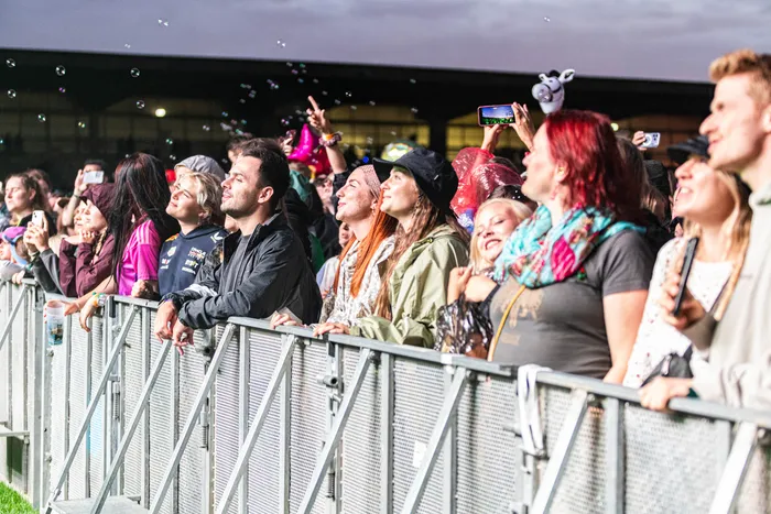 Fans stehen hinter einer Metallabsperrung bei einem Musikfestival und blicken in Richtung der Bühne, einige halten ihre Smartphones hoch.