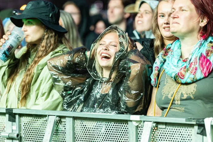 Eine lachende Frau in einem durchsichtigen Regenmantel steht in einer Menschenmenge hinter einer Metallabsperrung bei einem Musikfestival.