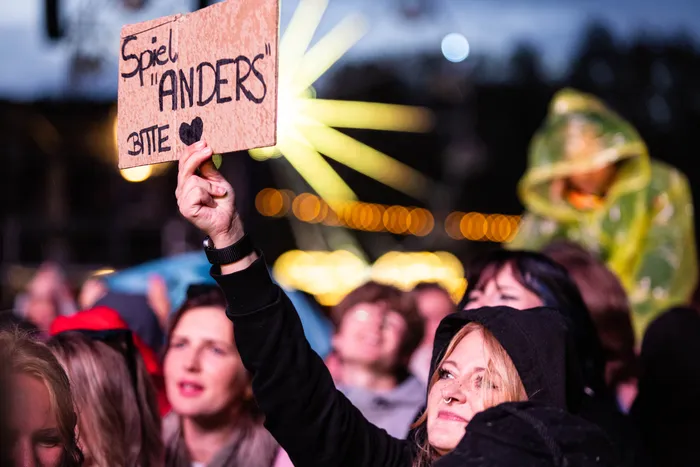 Eine Person hält ein Pappschild mit der Aufschrift „Spiel 'ANDERS' BITTE“ hoch in einer Menschenmenge bei einem Musikfestival.