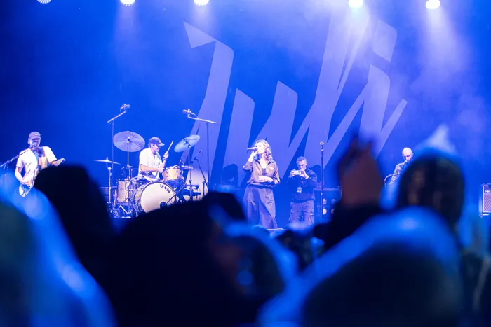 Die Band Juli tritt unter blauem Bühnenlicht auf einer Bühne auf. Im Hintergrund ist das große Bandlogo zu sehen, im Vordergrund das Publikum.