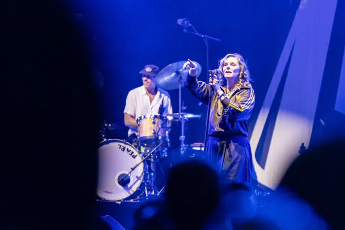 Sängerin der Band Juli singt unter blauem Bühnenlicht und zeigt mit einer Hand nach oben, während im Hintergrund ein Schlagzeuger spielt.