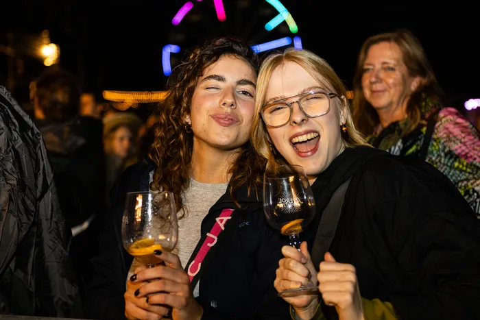 Zwei junge Frauen lachen und halten Weingläser in der Hand vor einem nächtlichen Hintergrund mit bunten Lichtern.