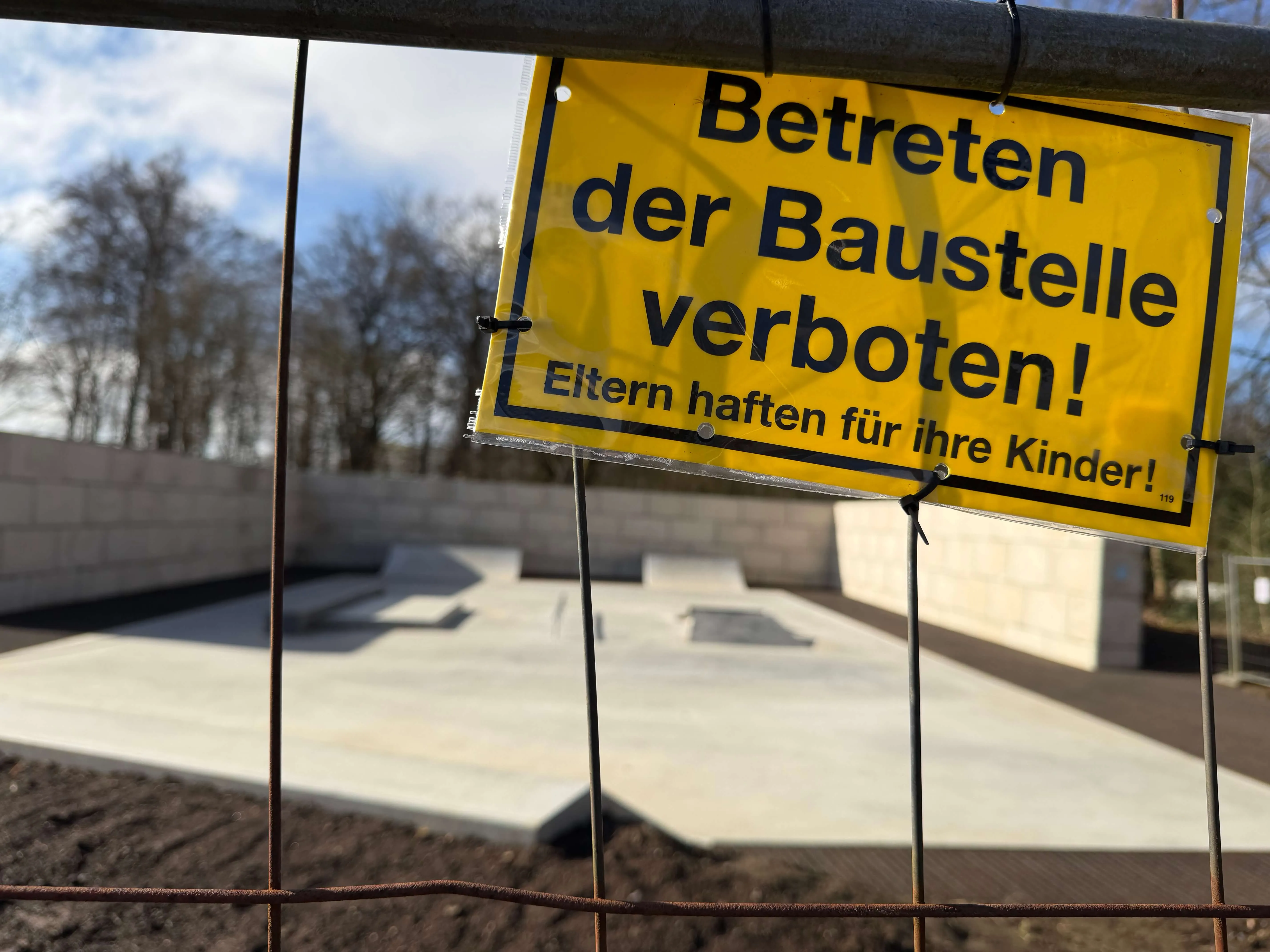Gelbes Warnschild mit der Aufschrift „Betreten der Baustelle verboten! Eltern haften für ihre Kinder!“ vor einer unfertigen Skateanlage.