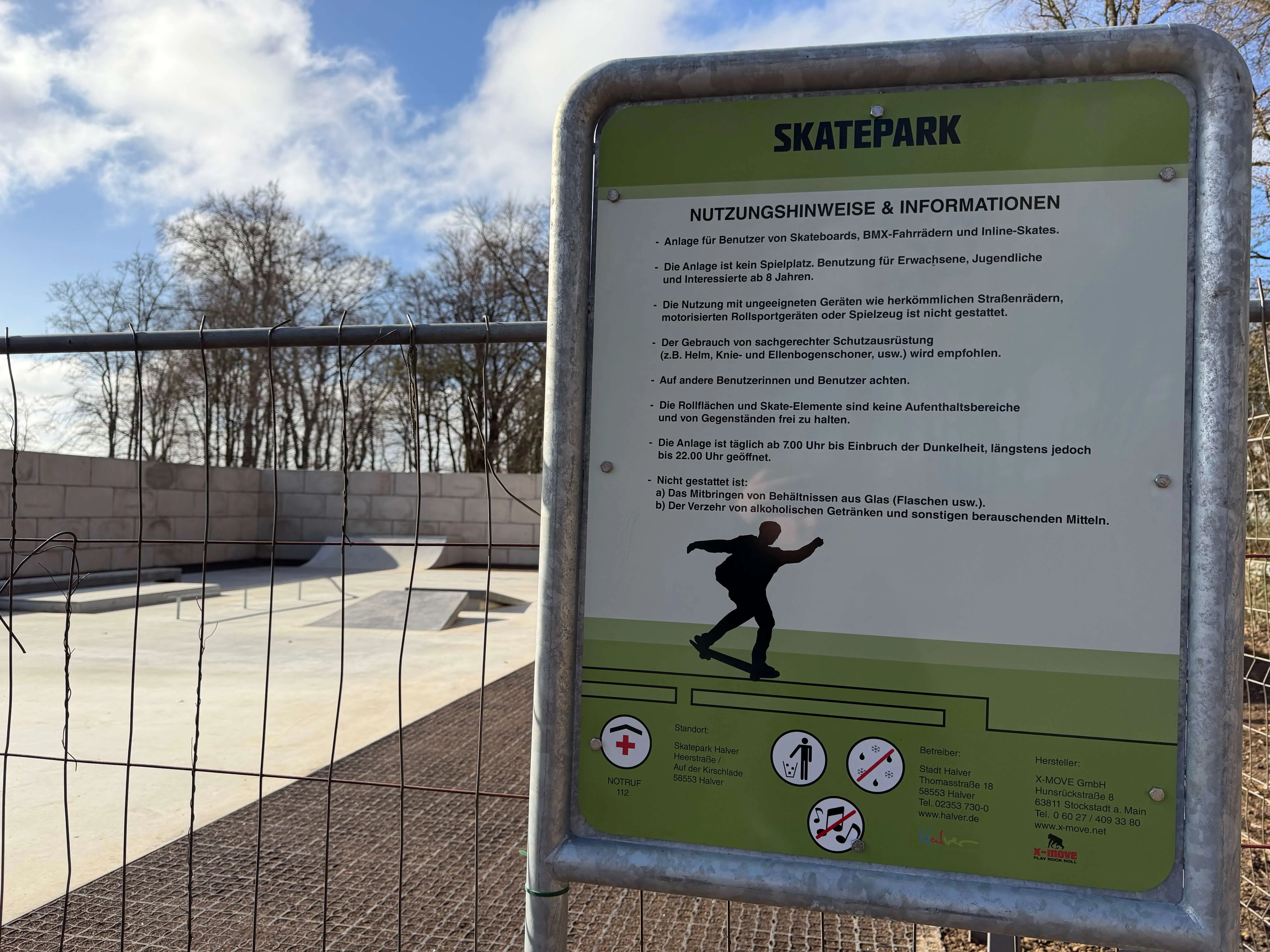 Schild mit Nutzungsordnung und Informationen vor einer Skateanlage, auf der ein Skater eine Rampe benutzt.