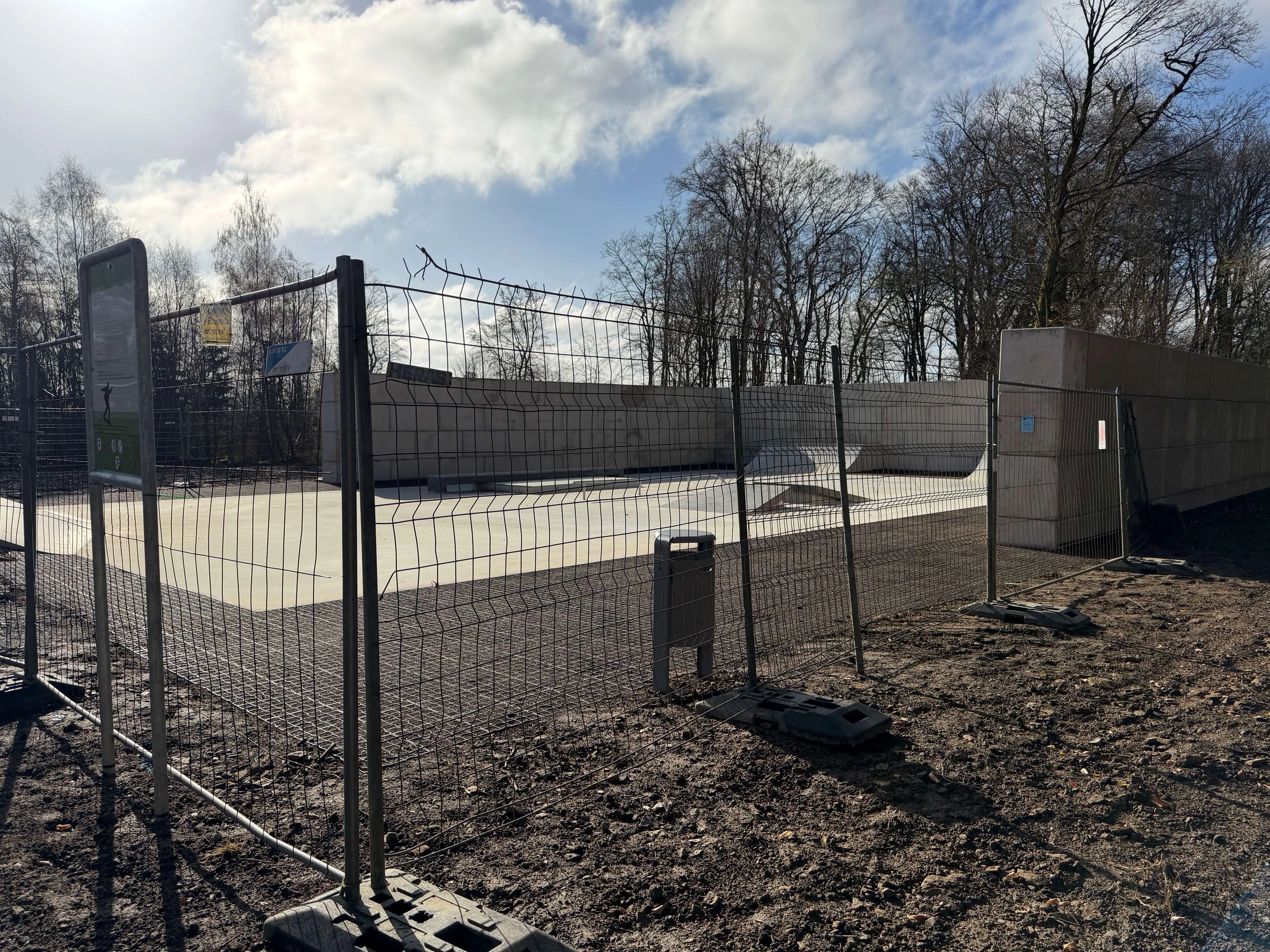 Betonkonstruktionen einer neuen Skate- und Bikeanlage sind durch einen Bauzaun abgetrennt. Im Hintergrund sind Bäume und ein bewölkter Himmel zu sehen.