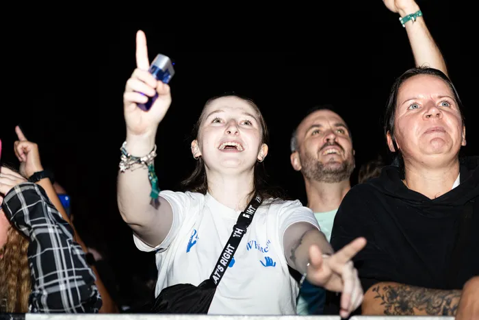 Eine lächelnde junge Frau in einem weißen T-Shirt zeigt mit ausgestrecktem Arm nach oben in einer Menschenmenge bei einem Festival.