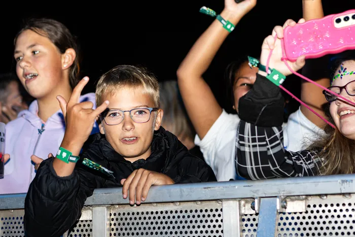 Ein Junge mit Brille macht eine Handgeste vor einer Metallabsperrung bei einem Festival.
