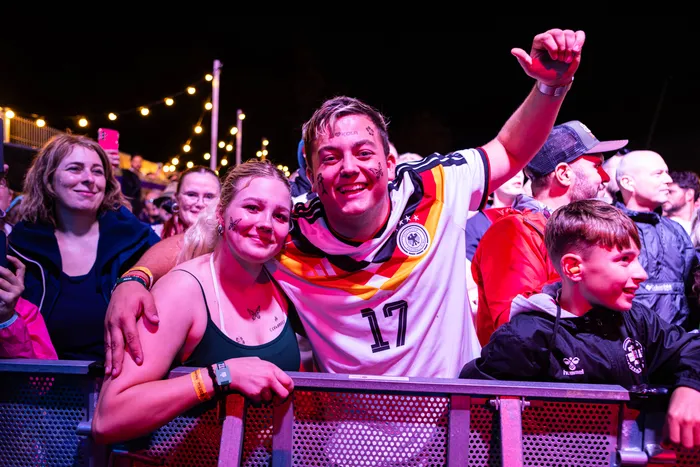 Ein junger Mann im deutschen Fußballtrikot und eine junge Frau mit Schmetterlings-Make-up lächeln hinter einer Absperrung bei einem nächtlichen Festival.