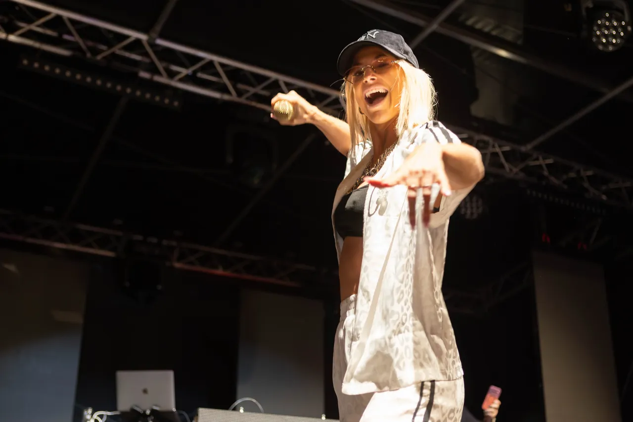 Isi Glück singt mit einem Mikrofon auf der Bühne beim Bautz Festival 2026. Sie trägt eine schwarze Kappe, eine Brille und ein weiß gemustertes Outfit.