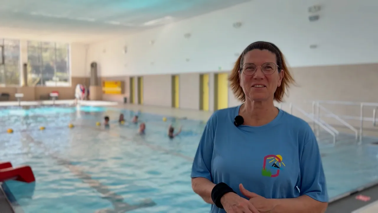 Lachende Frau mit Brille und blauem T-Shirt vor einem Schwimmbecken, in dem sich mehrere Personen befinden.