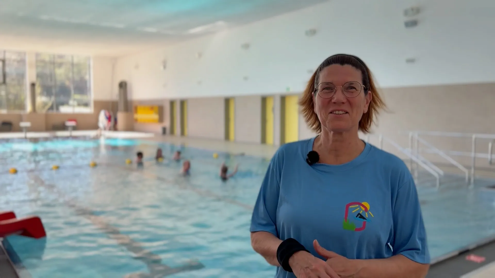 Lachende Frau mit Brille und blauem T-Shirt vor einem Schwimmbecken, in dem sich mehrere Personen befinden.