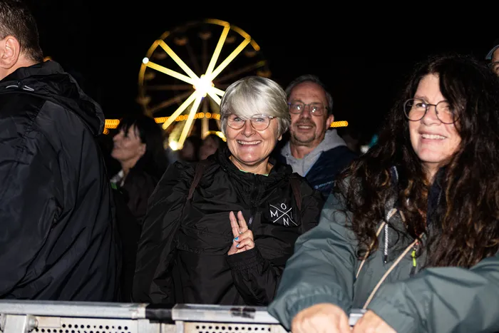 Eine lächelnde Frau mit kurzen grauen Haaren und Brille zeigt das Peace-Zeichen vor einem beleuchteten Riesenrad bei Nacht.