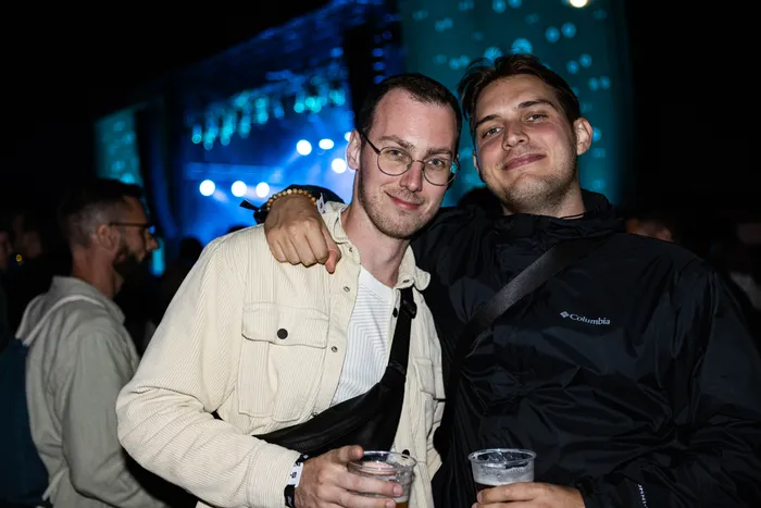 Zwei Männer lächeln in die Kamera und halten Getränke in Plastikbechern vor einer nächtlichen Festival-Kulisse.