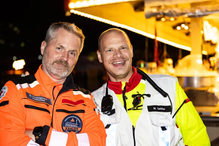 Zwei lächelnde Männer in Uniformen des Rettungsdienstes und Katastrophenschutzes vor einer hell erleuchteten Kulisse beim Bautz-Festival.