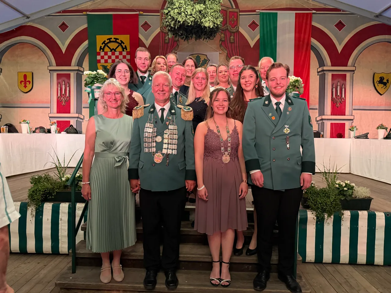 Eine Gruppe von Personen in festlicher Kleidung und grünen Schützenuniformen mit Orden posiert auf einer Bühne beim Schützenfest Hülscheid.