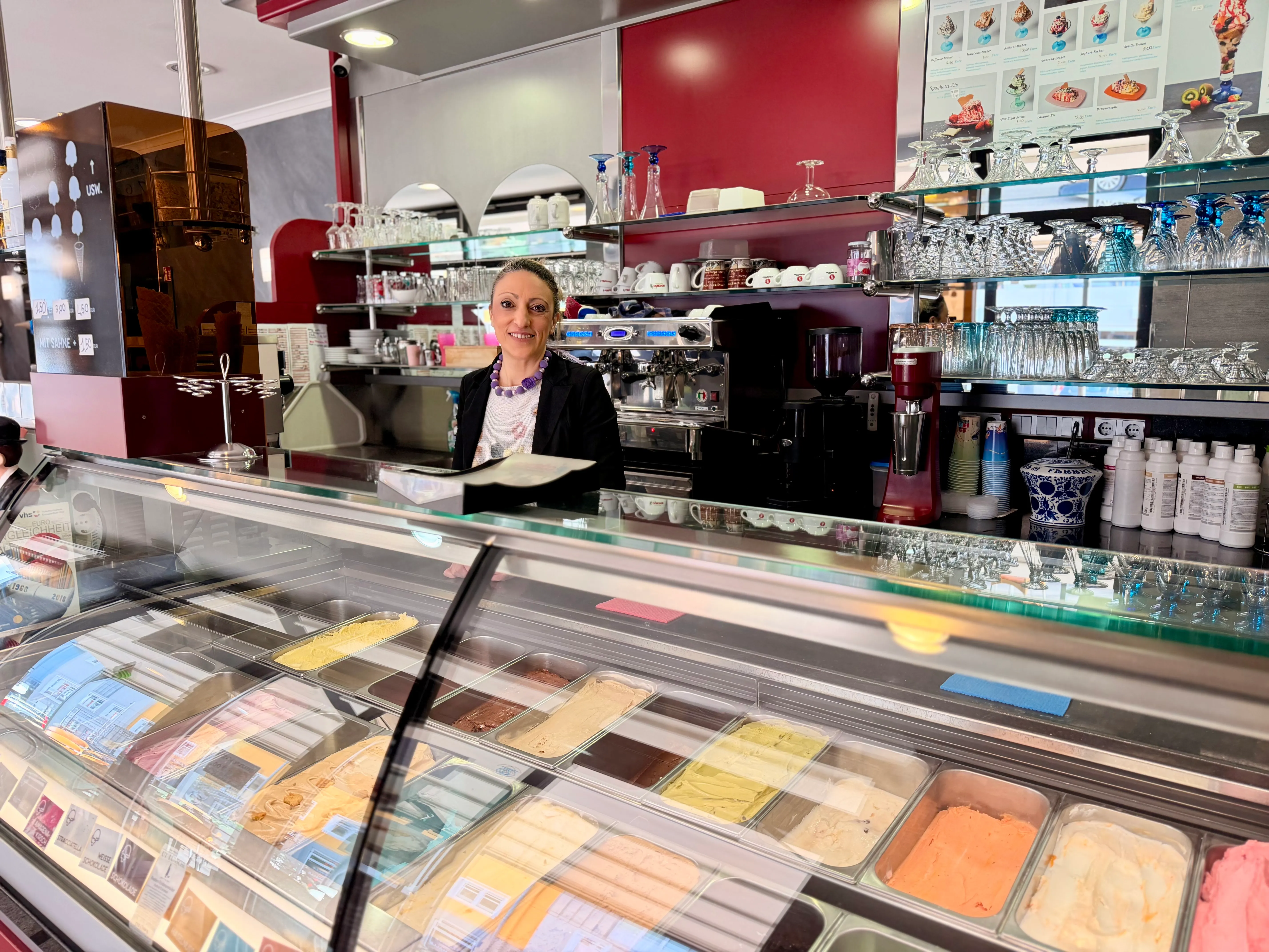 Eine Frau lächelt hinter der Theke einer Eisdiele. Verschiedene Eissorten sind in Kühlvitrinen zu sehen, im Hintergrund stehen Gläser und eine Kasse.