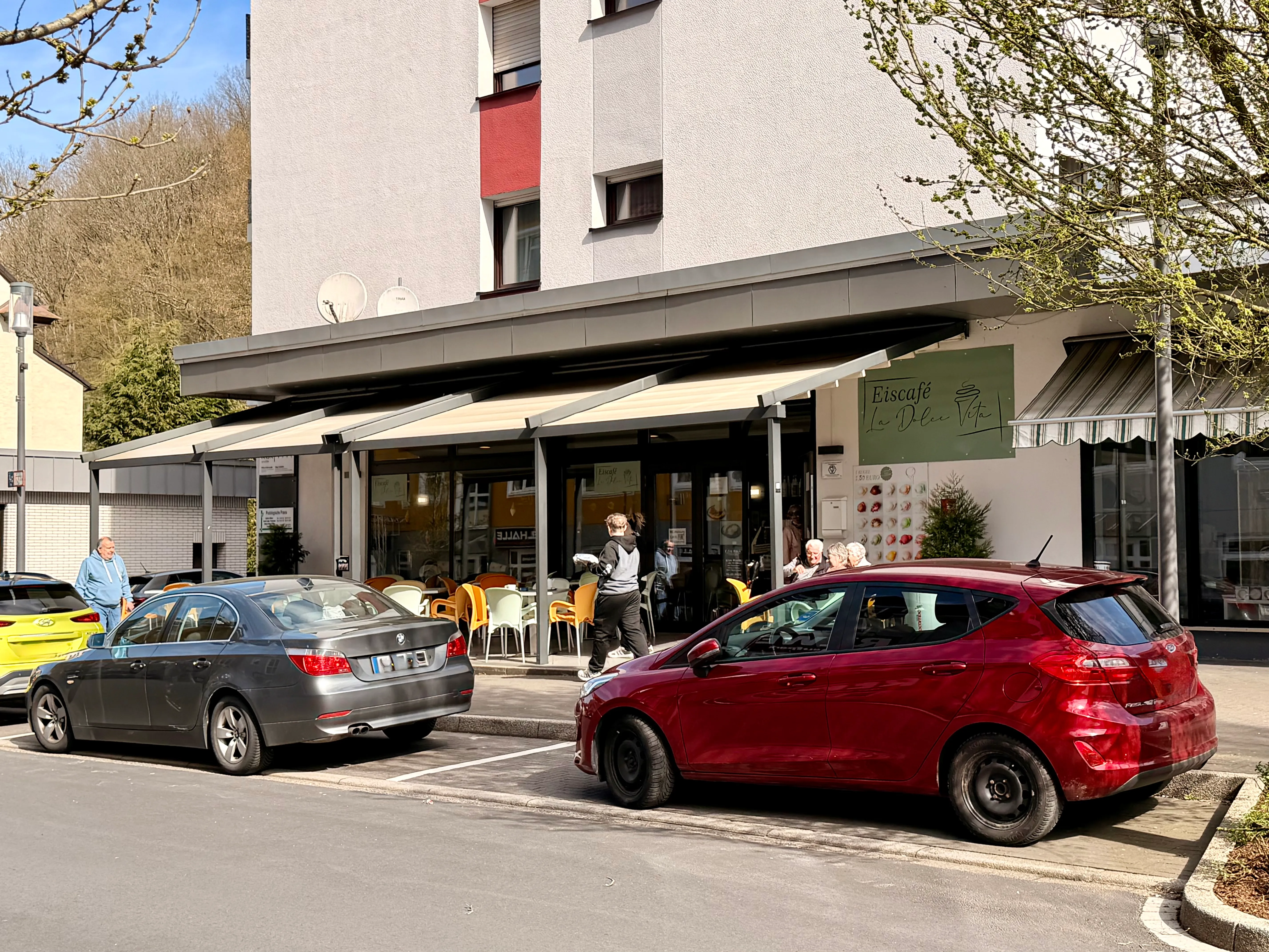 Eisdiele „La dolce vita“ mit einigen Gästen und parkenden Autos davor. Im Hintergrund ein Wohnhaus mit Satellitenschüssel.