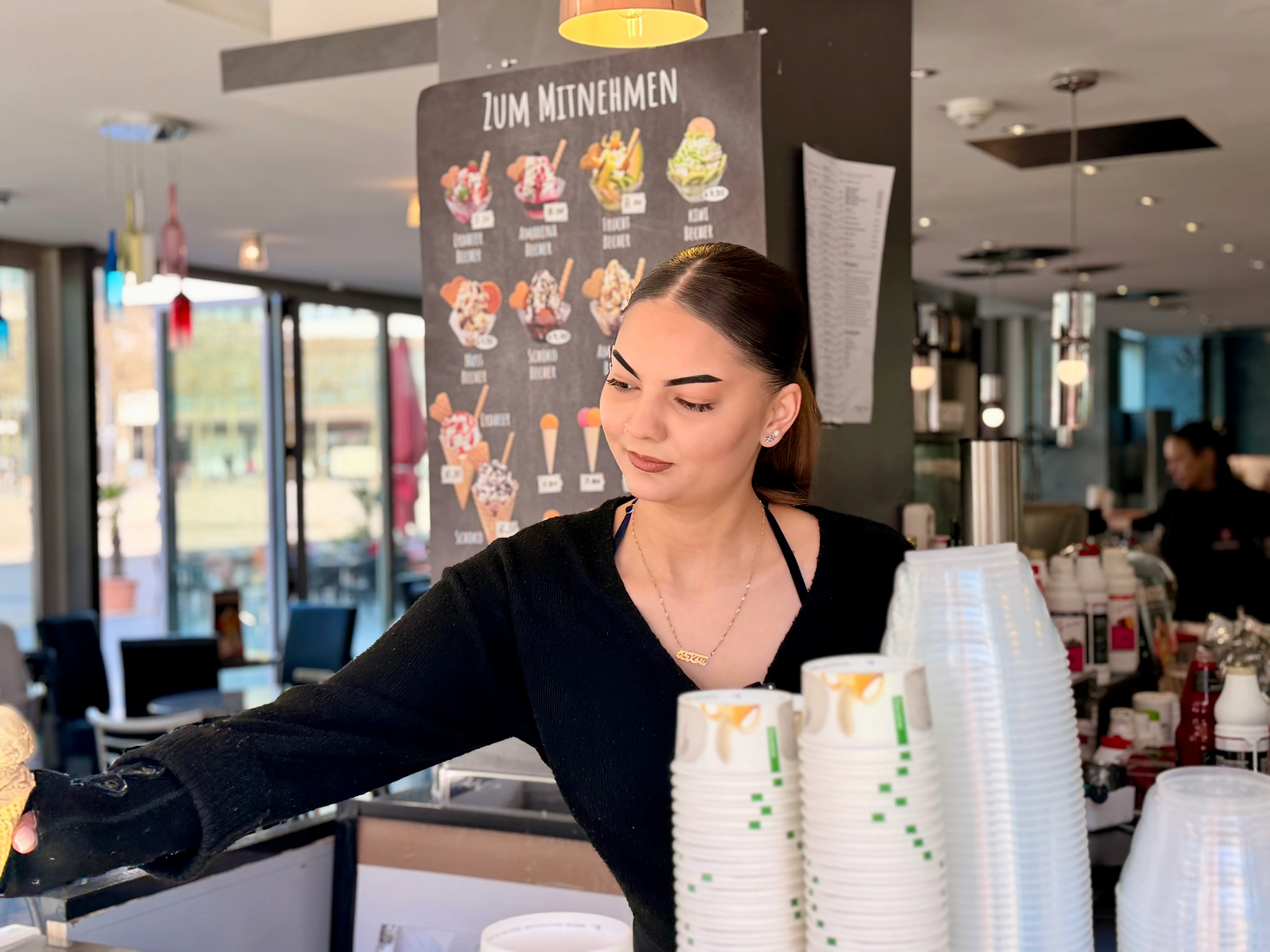 Junge Frau mit dunklen Hauren bedient im Eiscafé „Zentro“ in Lüdenscheid und reicht Becher über den Tresen. Im Hintergrund eine Menütafel mit verschiedenen Eissorten.