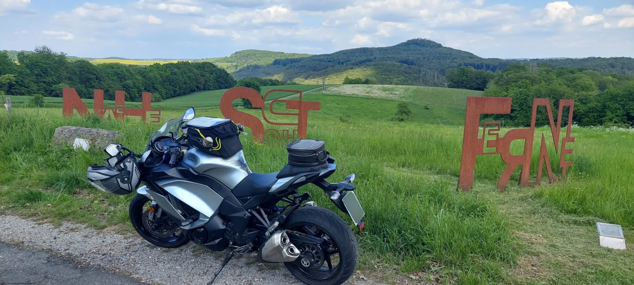 Motorrad steht auf einem Feldweg vor einer Hügellandschaft mit großen, roten Buchstaben, die ein Wort formen.