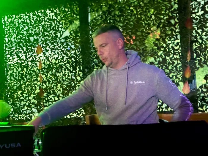 Ein Mann mit kurzgeschorenen Haaren und einem grauen Kapuzenpullover steht hinter einem Mischpult und bedient es. Im Hintergrund eine grüne Wand mit floralem Muster und Lichteffekte.