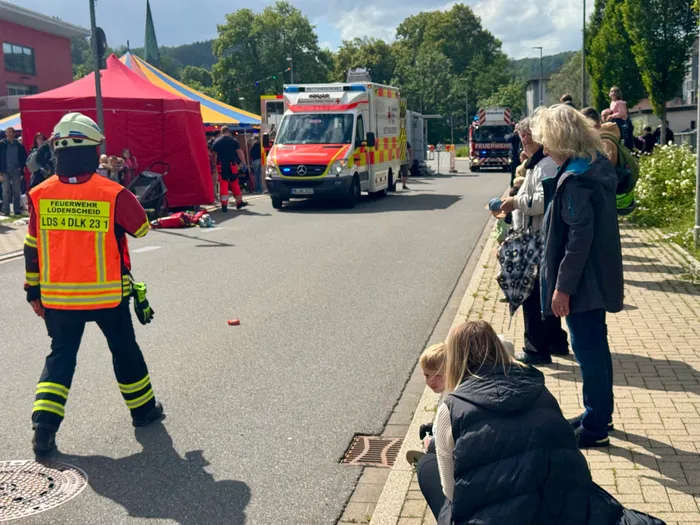 Feuerwehrmann in Warnweste steht vor einem Rettungswagen und weiteren Einsatzfahrzeugen. Im Hintergrund beobachten Menschen das Einsatzszenario.