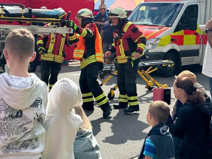Feuerwehrleute tragen eine Trage mit einem unbekannten Patienten an einem sonnigen Tag. Im Hintergrund sind Einsatzfahrzeuge und Zuschauer zu sehen.