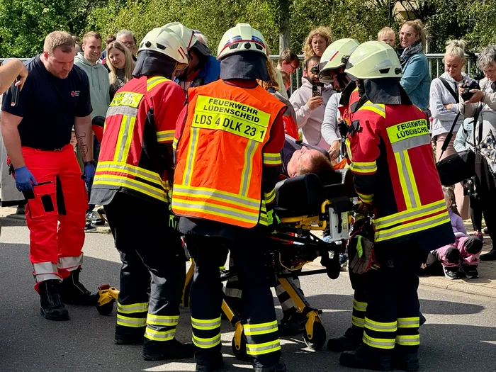 Feuerwehrleute in orangefarbenen Westen und Helmen transportieren eine Person auf einer Trage, umgeben von Einsatzkräften und Zuschauern. Auf den Westen ist 