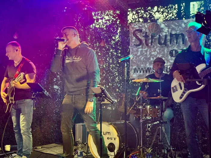 Eine Band spielt auf einer Bühne vor Publikum. Ein Sänger mit Kopfhörer steht im Vordergrund, während ein Gitarrist, ein Bassist und ein Schlagzeuger im Hintergrund zu sehen sind. Im Hintergrund befindet sich ein Banner mit der Aufschrift „Strum“ und einer Website-Adresse.