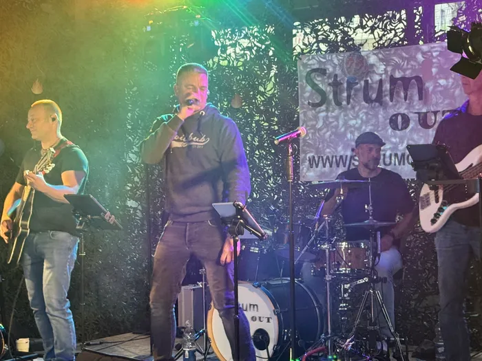 Ein Sänger mit Mikrofon steht auf einer Bühne vor einer Band. Im Hintergrund ist ein Banner mit der Aufschrift „Strum Out“ und einer Webseite zu sehen.