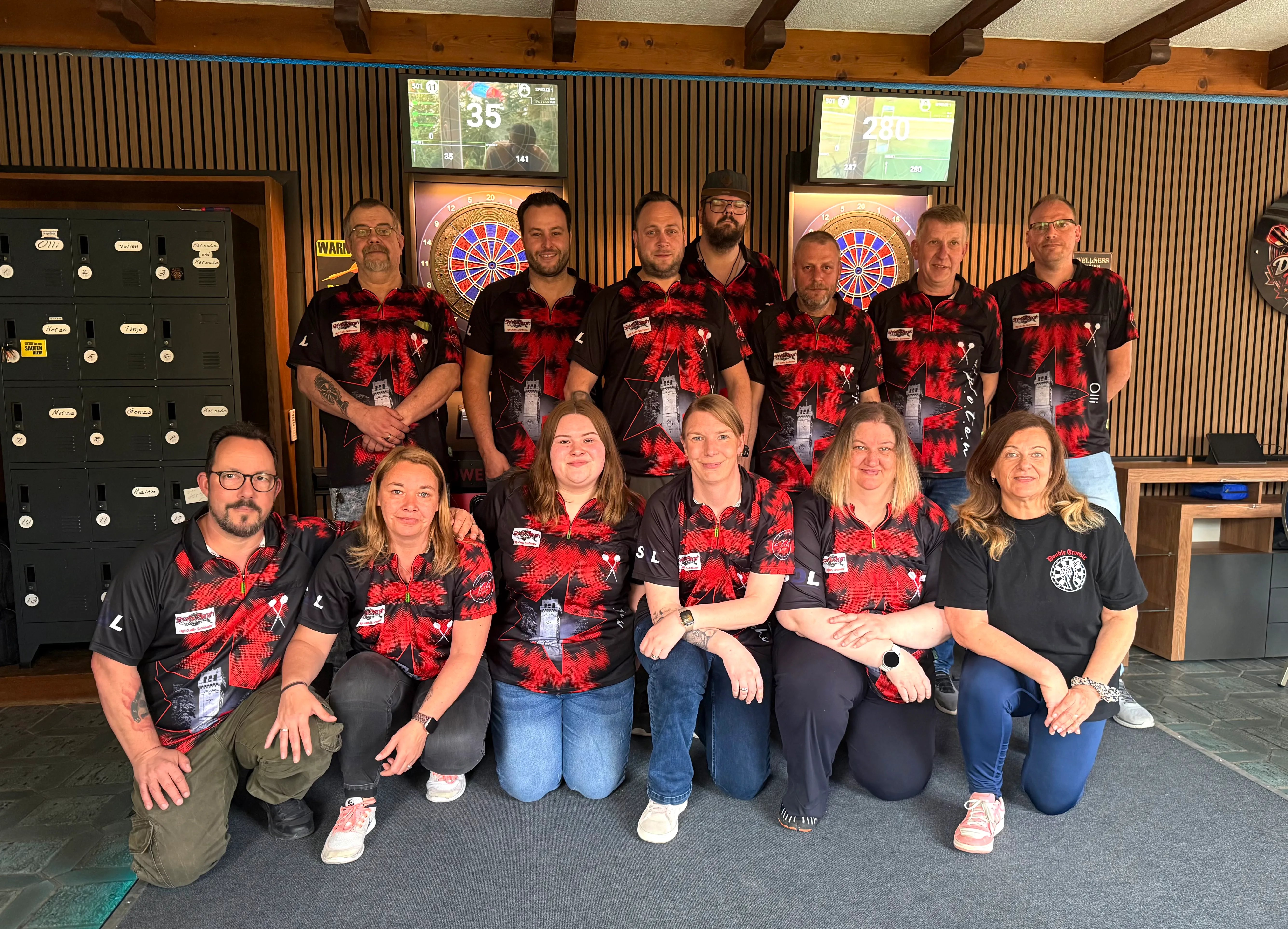 Gruppe von etwa zwanzig Dartsspielern in Vereinskleidung posiert vor einer Dartscheibe und elektronischen Anzeigetafeln im neuen Vereinsheim.