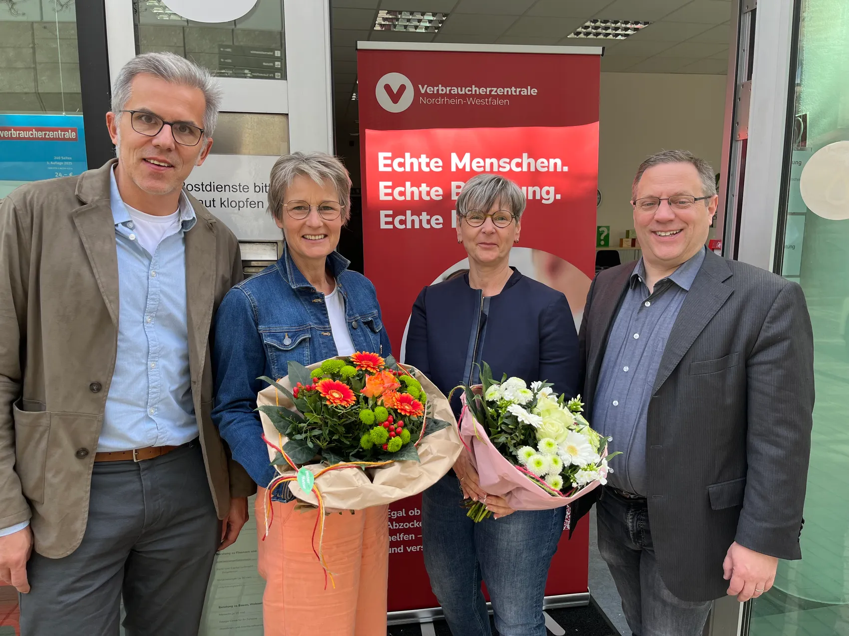 Vier Personen vor dem Eingang der Verbraucherzentrale Nordrhein-Westfalen in Lüdenscheid, zwei Frauen halten Blumensträuße.