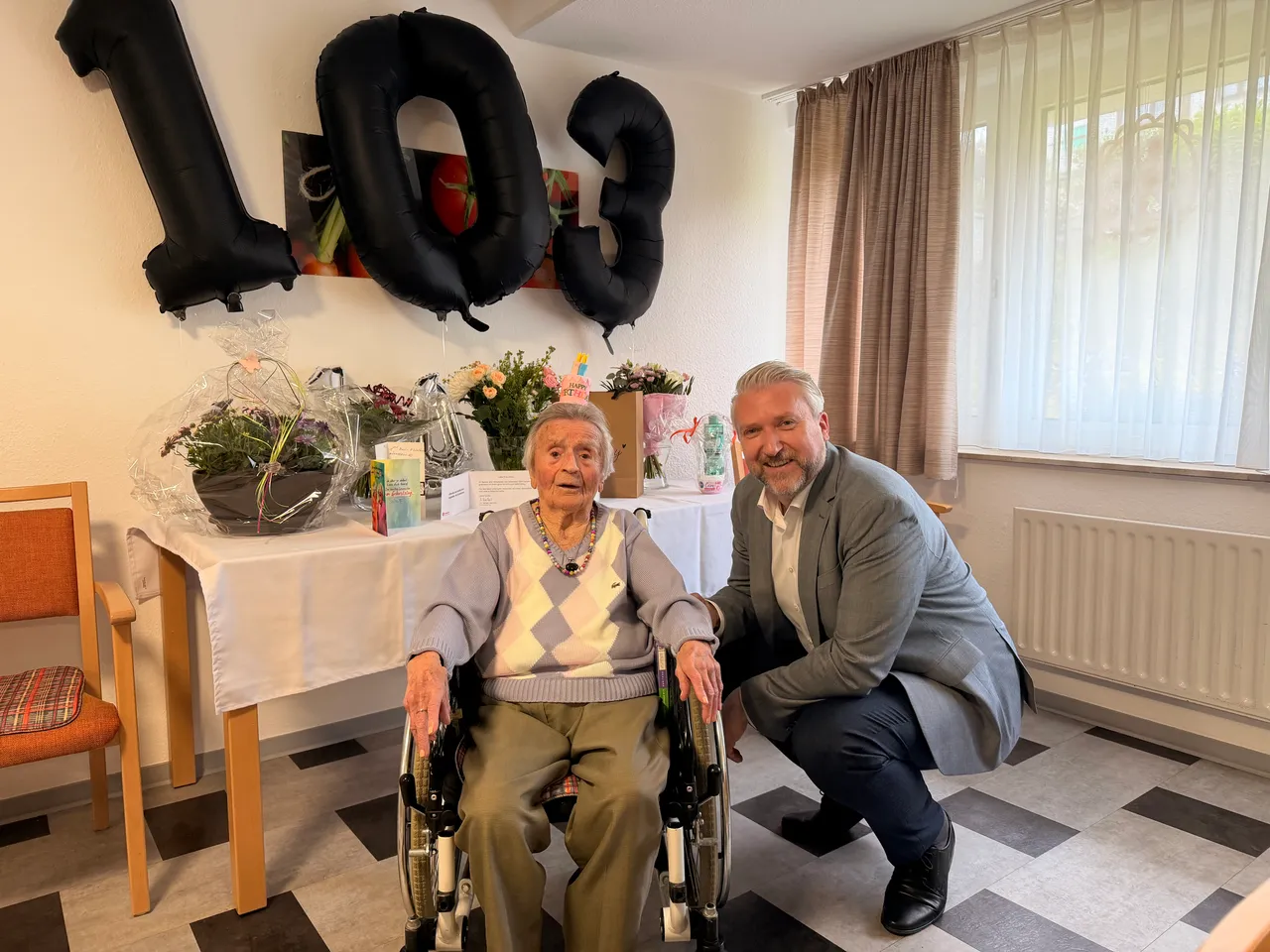Eine ältere Frau im Rollstuhl lächelt neben einem Mann in einem hellen Raum, im Hintergrund große schwarze Ballons mit der Zahl 103.