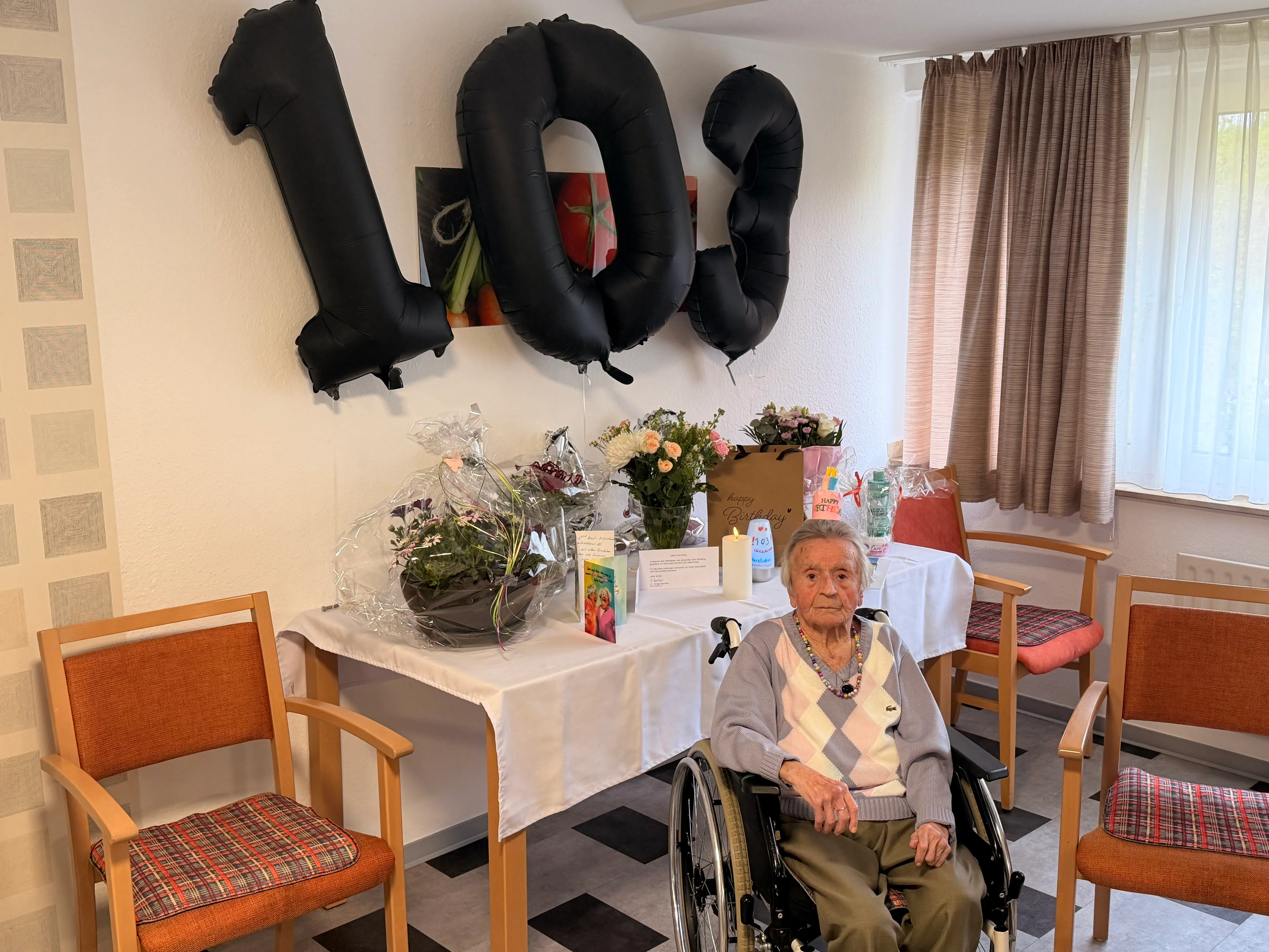 Eine ältere Frau sitzt im Rollstuhl vor einem Tisch mit Blumen, Geschenken und schwarzen Folienballons in Form der Zahlen 1 und 0.