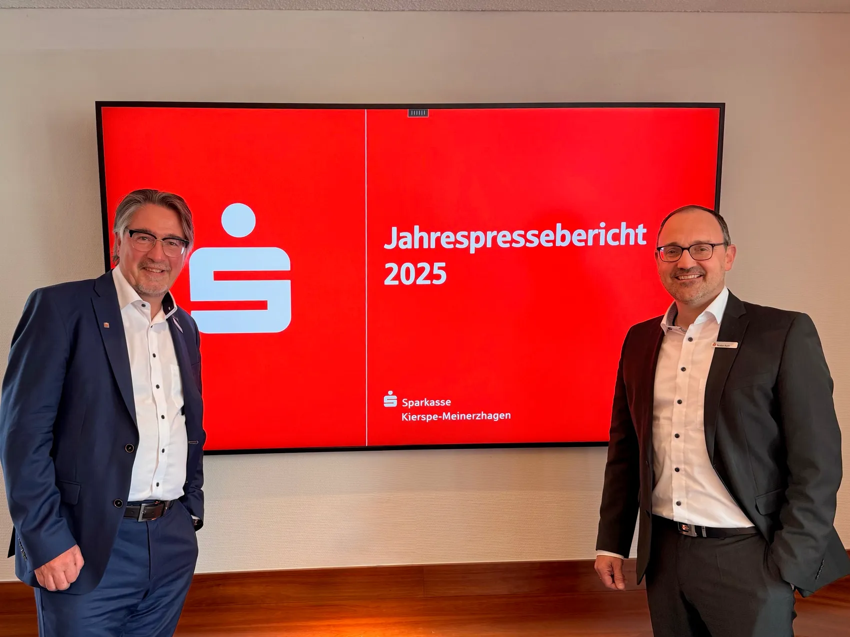 Zwei Männer im Anzug stehen vor einer Präsentation des Jahrespresseberichts 2025 der Sparkasse Kierspe-Meinerzhagen.