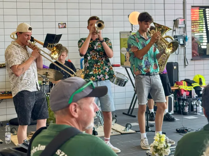 Vier junge Männer spielen Posaune und Tenorhorn auf einer kleinen Bühne. Im Hintergrund sind weitere Personen und Gegenstände in einem hellen Raum zu sehen.