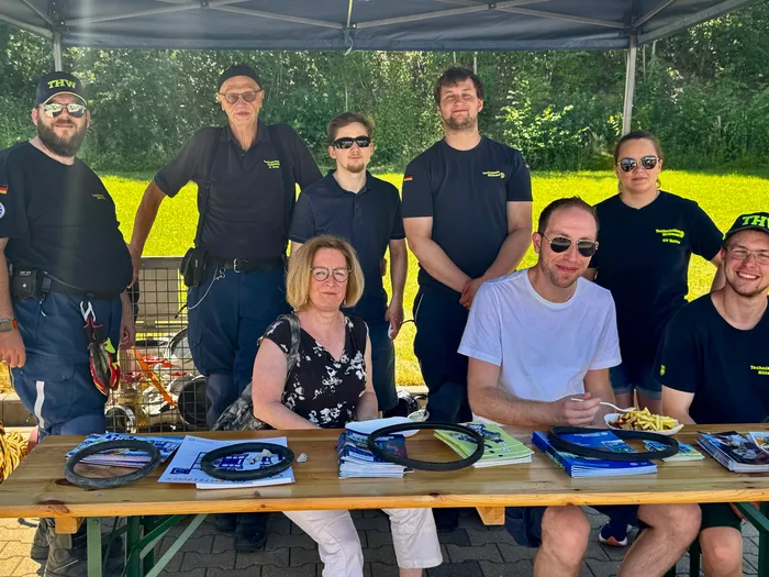 Eine Gruppe von Männern und Frauen, die meisten in Uniformen des Technischen Hilfswerks (THW), posieren vor einem Tisch mit Informationsmaterialien und Speisen. Im Hintergrund steht ein Pavillon auf einer grünen Wiese.