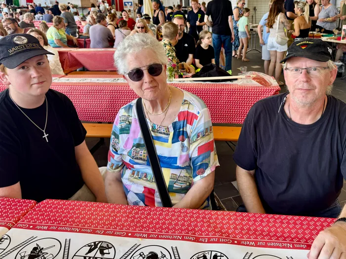 Drei Generationen sitzen an einem Tisch mit roter und weißer karierter Tischdecke. Im Hintergrund sind Menschen und ein Festzelt zu sehen.