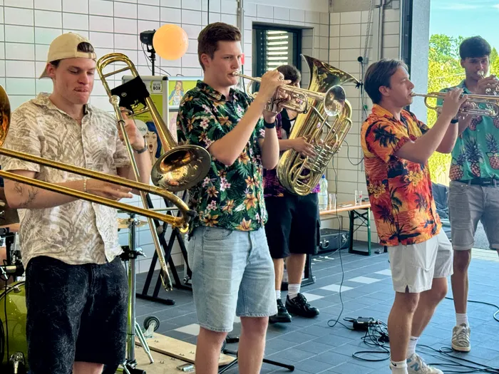 Eine Bläsergruppe spielt auf einem öffentlichen Platz. Vier Musiker mit Posaune und Trompete sind im Vordergrund zu sehen, während weitere Musiker im Hintergrund stehen.