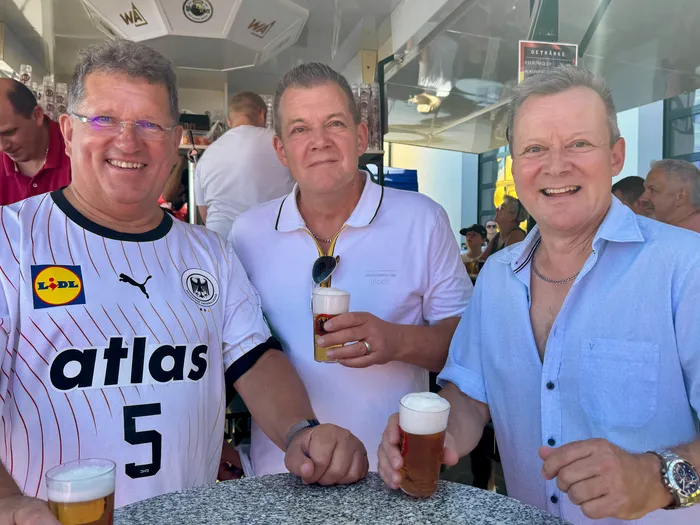 Drei Männer lächeln in die Kamera, jeder hält ein Bierglas oder eine Bierflasche. Der Mann links trägt ein Fußballtrikot, der Mann in der Mitte ein weißes Poloshirt und der Mann rechts ein blaues Hemd.