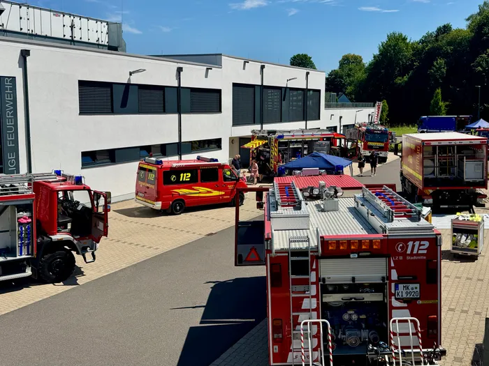 Mehrere rote und orangefarbene Feuerwehrautos stehen auf dem Hof einer Feuerwache, umgeben von Feuerwehrleuten und Besuchern. Im Hintergrund ein modernes Gebäude mit großen Fenstern und Bäumen.