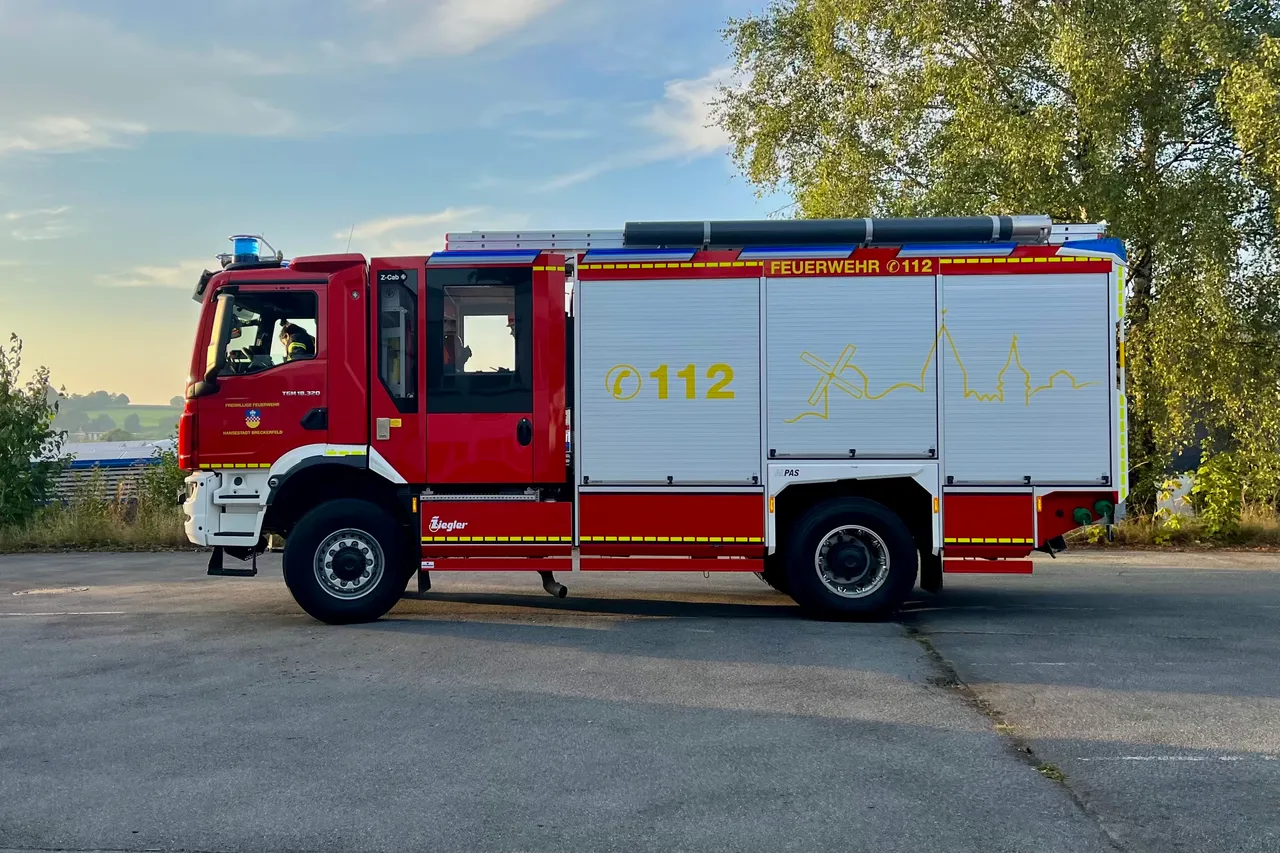 Roter Feuerwehrauto mit der Aufschrift „Feuerwehr 112“ und einem stilisierten Flammenmotiv an der Seite, geparkt an einer Straße mit Bäumen im Hintergrund.